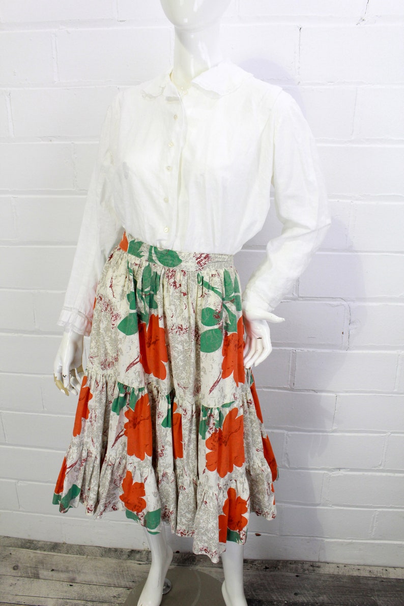 May include: A white button-up shirt with a peter pan collar paired with a tiered skirt featuring a floral print in shades of orange, green, and beige.