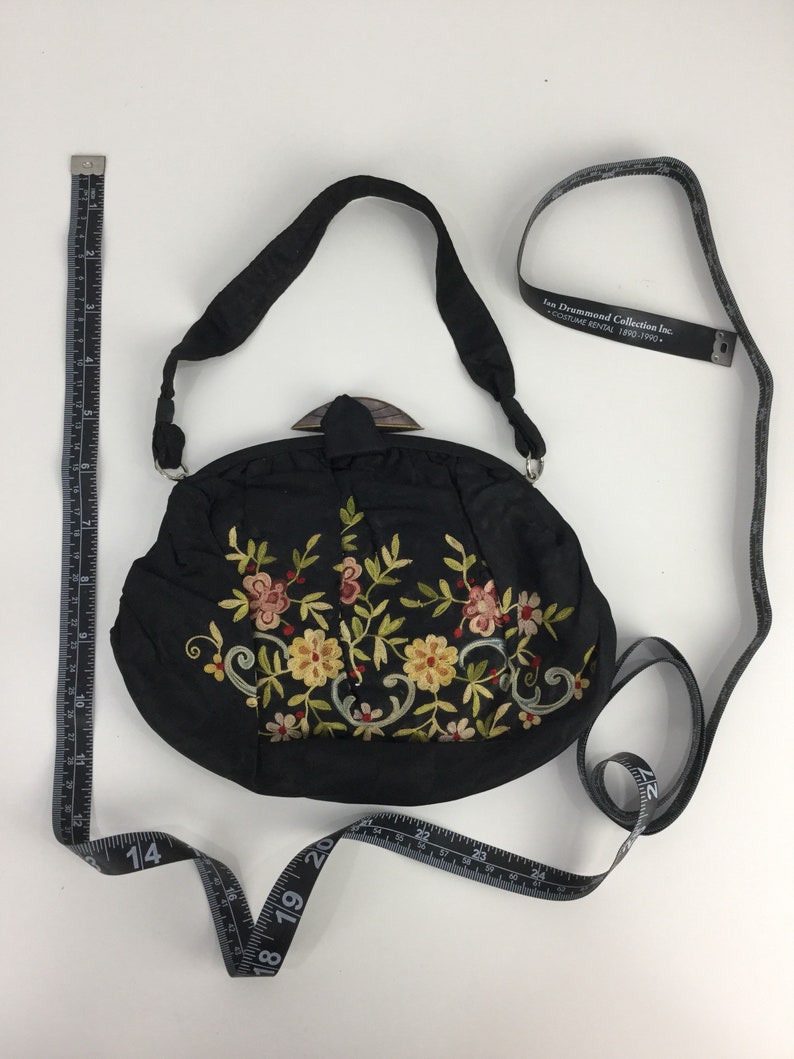 Pode incluir: Bolsa de noite de cetim preto bordada com um design floral e um fecho de madeira. A bolsa tem uma al&ccedil;a comprida e mede 18 cm de di&acirc;metro.
