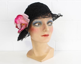 May include: A black lace cloche hat with a pink flower accent. The hat is on a mannequin head.