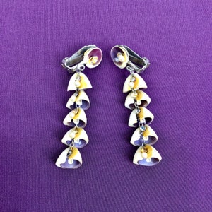 May include: A pair of long, dangling earrings made of seashells. Each earring has six seashells, each with a pearl-like bead in the center. The seashells are a mix of purple, white, and yellow.