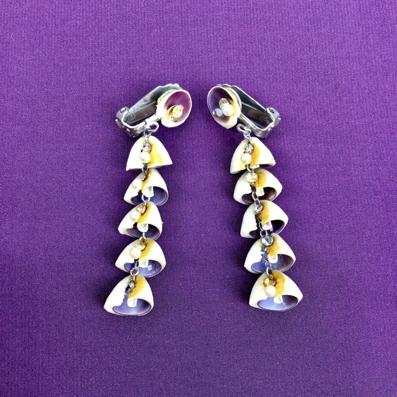 May include: A pair of long, dangling earrings made of seashells. Each earring has six seashells, each with a pearl-like bead in the center. The seashells are a mix of purple, white, and yellow.