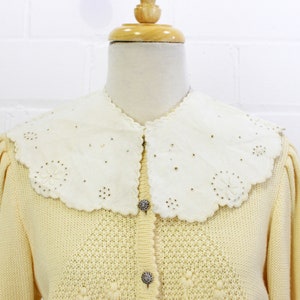 May include: A pale yellow knitted cardigan with a white lace collar and silver buttons. The cardigan has a delicate floral pattern.