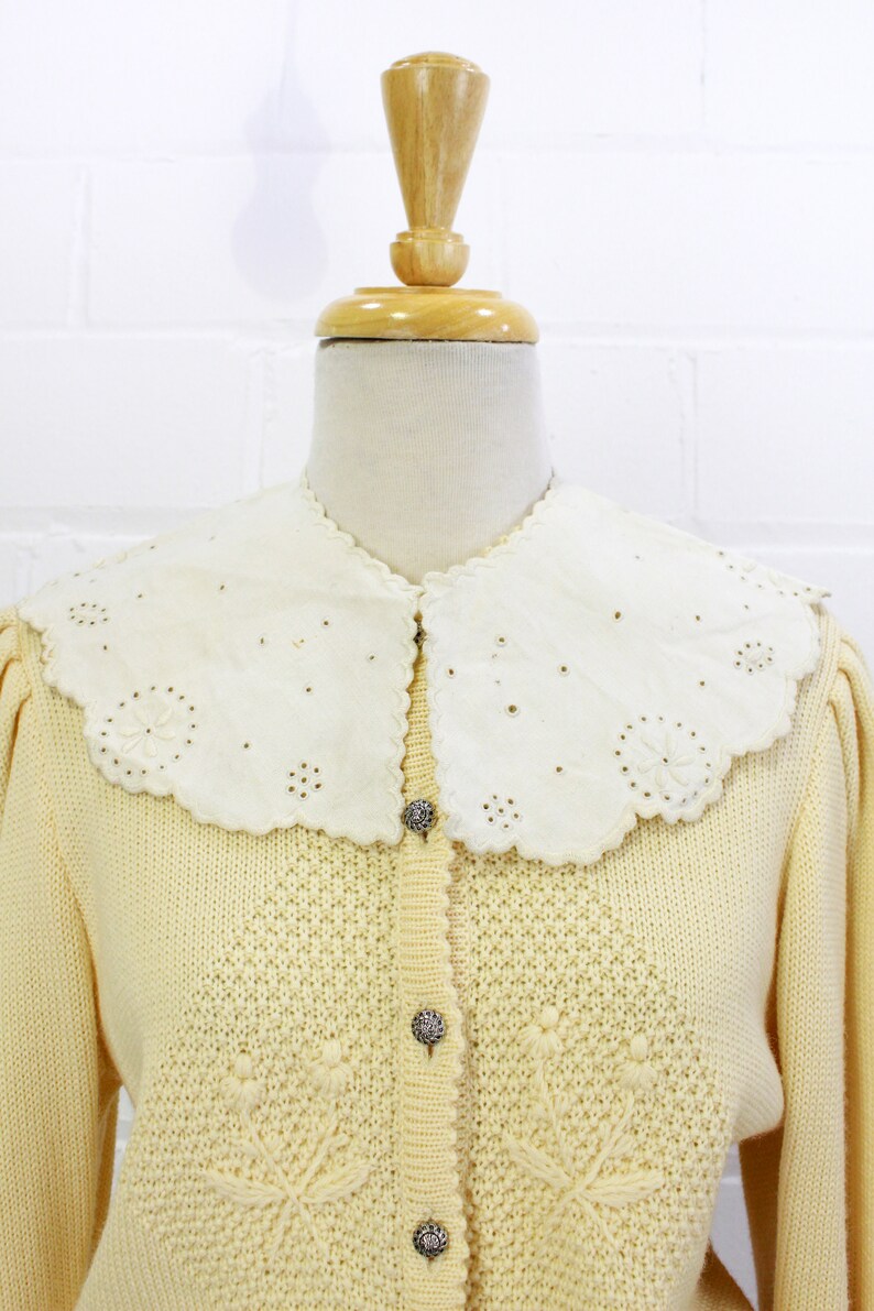 May include: A pale yellow knitted cardigan with a white lace collar and silver buttons. The cardigan has a delicate floral pattern.