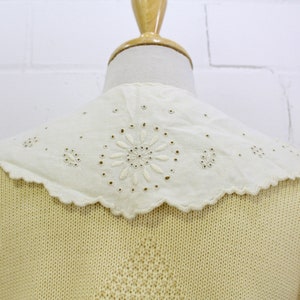 May include: A pale yellow knitted cardigan with a white eyelet lace collar. The collar has a scalloped edge and a delicate floral design.