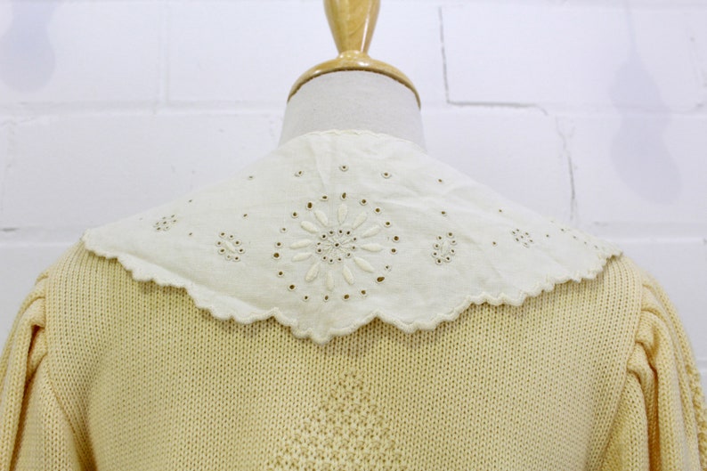 May include: A pale yellow knitted cardigan with a white eyelet lace collar. The collar has a scalloped edge and a delicate floral design.