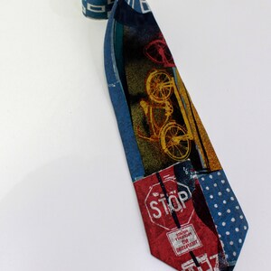 May include: A patterned necktie with a bicycle and stop sign design. The tie features a mix of blue, red, and yellow colors. The tie is rolled up at the top and lies on a white surface.