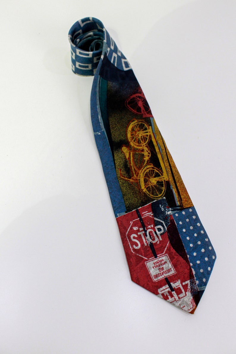 May include: A patterned necktie with a bicycle and stop sign design. The tie features a mix of blue, red, and yellow colors. The tie is rolled up at the top and lies on a white surface.
