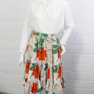 May include: A white long-sleeved button-up shirt with a peter pan collar paired with a tiered skirt featuring a floral print in shades of orange, green, and white.