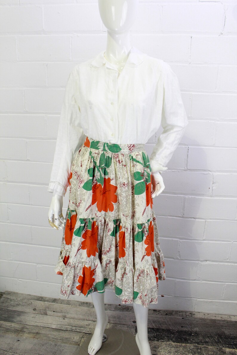 May include: A white long-sleeved button-up shirt with a peter pan collar paired with a tiered skirt featuring a floral print in shades of orange, green, and white.
