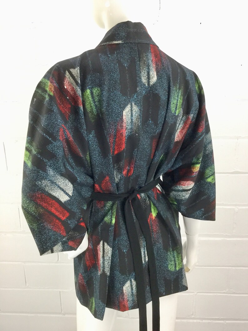 May include: A black kimono with a colorful abstract print featuring red, green, and white brushstrokes. The kimono has a black sash.