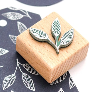 May include: A wooden stamp featuring a green leaf design. The stamp is square and has a light wood finish.