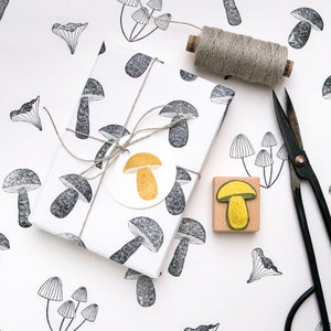 May include: A white gift wrap with a black and white mushroom pattern. The gift is tied with twine and a yellow mushroom sticker. A wooden stamp with a yellow mushroom design and a pair of black scissors are also shown.