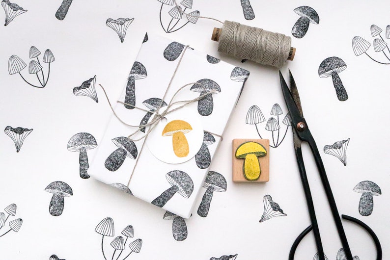 May include: A white gift wrap with a black and white mushroom pattern. The gift is tied with twine and a yellow mushroom sticker. A wooden stamp with a yellow mushroom design and a pair of black scissors are also shown.