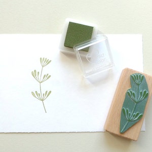 K&ouml;nnte beinhalten: Ein gr&uuml;nes Stempelkissen und ein Stempel aus Holz mit einem gr&uuml;nen Blattdesign. Der Stempel ist mit "tsa color" beschriftet.