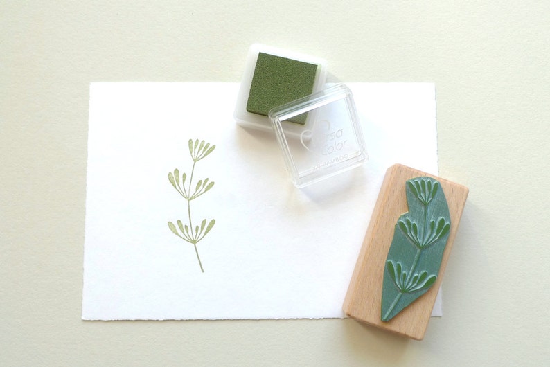 K&ouml;nnte beinhalten: Ein gr&uuml;nes Stempelkissen und ein Stempel aus Holz mit einem gr&uuml;nen Blattdesign. Der Stempel ist mit "tsa color" beschriftet.