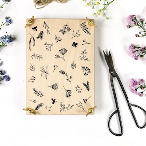 May include: A wooden block with botanical illustrations, secured by brass fixings, is surrounded by sprigs of blue and pink flowers, twine, and vintage scissors. The block features various hand-drawn floral and leaf designs.