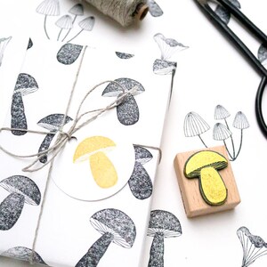 May include: A white gift box with a black and white mushroom pattern. The box is tied with twine and has a round, yellow mushroom sticker. A wooden stamp with a yellow mushroom design is also shown.