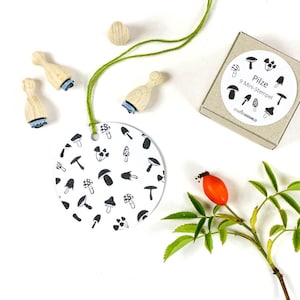 Può includere: Una collezione di articoli per l'artigianato a tema funghi. Include timbri in legno, un'etichetta con un motivo a funghi e una scatola con la scritta "Pilze 9 Mini-Stempel". Sono presenti anche un rametto verde e una bacca rossa.