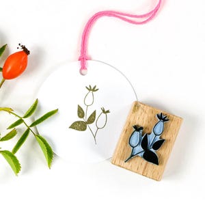 Op de afbeelding: Een witte, ronde tag met een gouden botanische illustratie en een roze touwtje, naast een houten stempel met een vergelijkbaar ontwerp. Een takje groene bladeren en een oranje rozenbottel zijn ook aanwezig.