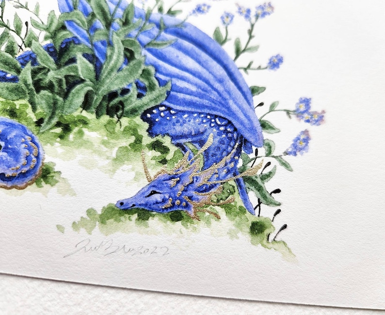 K&ouml;nnte beinhalten: Ein Aquarellbild eines blauen Drachen mit goldenen Akzenten, der sich in einem Bett aus gr&uuml;nen Bl&auml;ttern und blauen Blumen zusammengerollt hat. Die Augen des Drachen sind geschlossen und er scheint zu schlafen. Das Bild ist unten rechts mit "Red 2022" signiert.