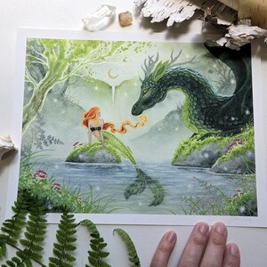 May include: A rectangular art print featuring a watercolor illustration of a mermaid with long orange hair sitting on a rock in a pond, facing a large dark green dragon with antlers. The scene includes a light green forest, a crescent moon, and small glowing lights.