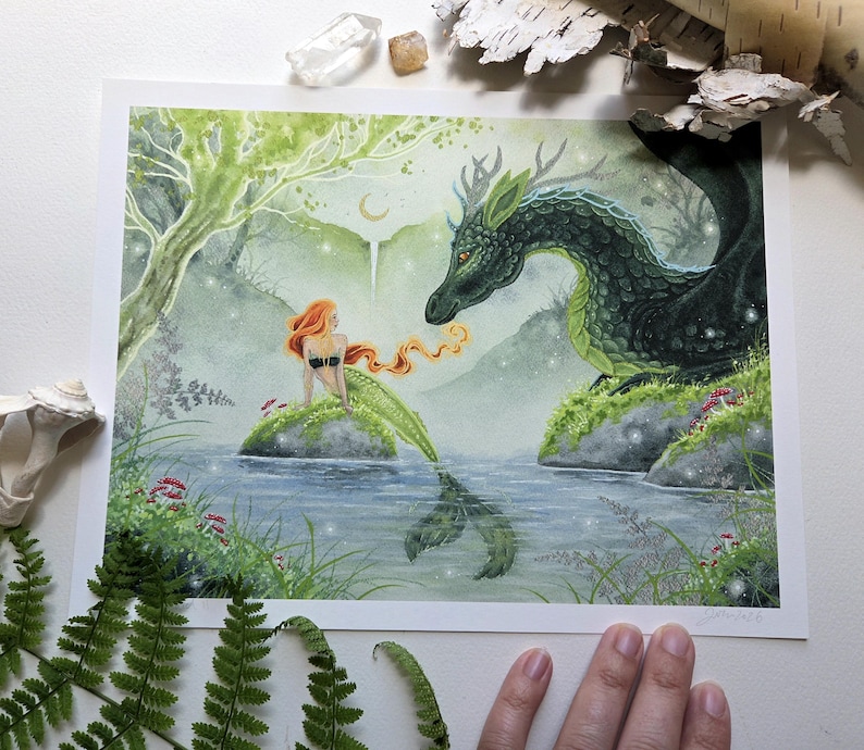 May include: A rectangular art print featuring a watercolor illustration of a mermaid with long orange hair sitting on a rock in a pond, facing a large dark green dragon with antlers. The scene includes a light green forest, a crescent moon, and small glowing lights.