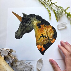 May include: A watercolor illustration of a unicorn head with a golden horn and black mane. The interior of the unicorn's head features a fall scene with a black unicorn, pumpkins, and a tree. The artwork is set against a white background.