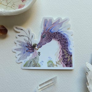 Pode incluir: Um autocolante com uma cena de fantasia de uma fada e um drag&atilde;o. O drag&atilde;o &eacute; principalmente roxo com detalhes rosa e dourado. A fada est&aacute; vestida com um vestido rosa e tem asas roxas. O fundo &eacute; branco.
