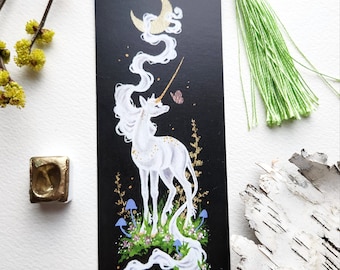 Puede incluir: Un marcapáginas negro con un unicornio blanco de pie en un campo de flores y setas bajo una luna creciente. La crin y la cola del unicornio ondean con el viento. El marcapáginas tiene un borla verde.
