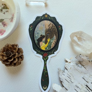 May include: A decorative sticker featuring a hand mirror design. The mirror's frame is dark green with floral accents, and the image inside depicts a woman in a yellow dress touching a dragon. A rose is at the base of the handle.