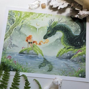 May include: A rectangular art print featuring a watercolor illustration of a mermaid with long orange hair sitting on a rock in a river, facing a large green dragon with antlers. The scene includes a forest background, a crescent moon, and small red mushrooms.