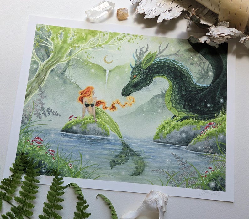 May include: A rectangular art print featuring a watercolor illustration of a mermaid with long orange hair sitting on a rock in a river, facing a large green dragon with antlers. The scene includes a forest background, a crescent moon, and small red mushrooms.