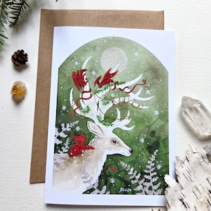 Könnte beinhalten: Aquarell-Grußkarte mit einem weißen Hirsch mit großem Geweih, verziert mit roten Akzenten und Grün. Der Hintergrund ist eine Mischung aus Grün und Weiß, mit einem Vollmond und fallenden Schneeflocken. Die Karte liegt auf einem braunen Umschlag.