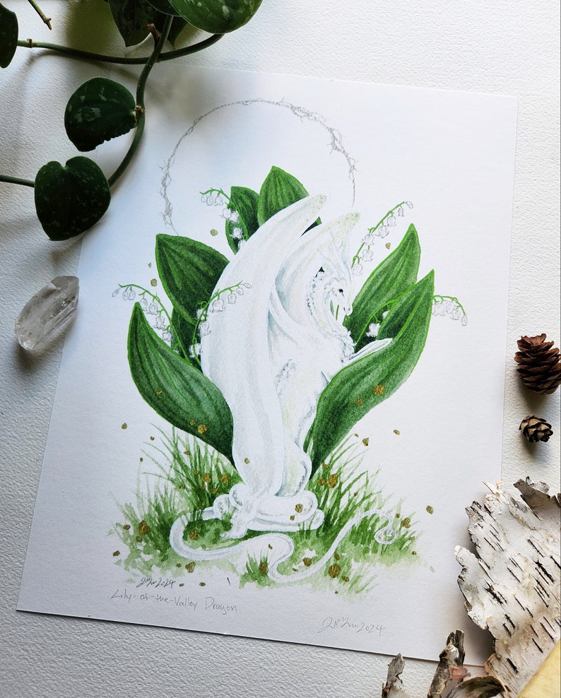 Puede incluir: Ilustraci&oacute;n en acuarela de un drag&oacute;n blanco emergiendo de hojas verdes y flores de lirios del valle. El drag&oacute;n tiene detalles intrincados y est&aacute; rodeado por un halo. La obra est&aacute; firmada y titulada "Lily-of-the-Valley Dragon".