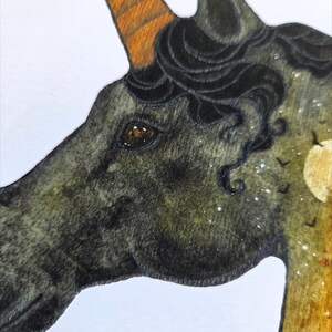 May include: A detailed illustration of a unicorn's head. The unicorn has a dark gray coat, a brown and orange horn, and black curly hair. The background features a moon, stars, and flying birds.