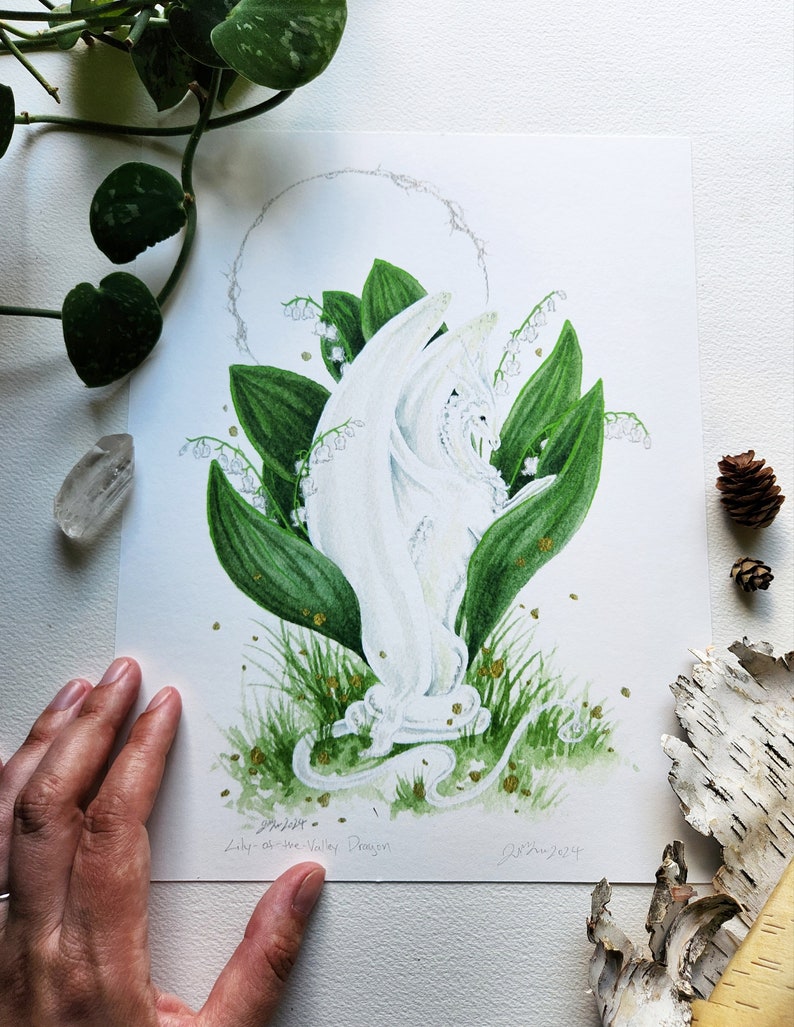 Puede incluir: Ilustraci&oacute;n en acuarela de un drag&oacute;n blanco entre hojas y flores verdes de lirios de los valles. La obra incluye motas doradas y un halo plateado. El texto "Lily-of-the-Valley Dragon" es visible.