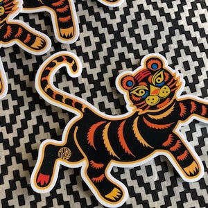 May include: A black and orange tiger sticker with a white outline. The tiger is standing on its hind legs with its front paws raised. The tiger has a yellow face with blue eyes and a red nose.