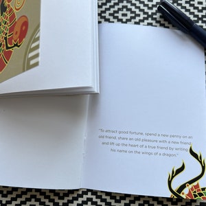 May include: A small, white book with a gold and red dragon design on the cover. The book has a quote on the inside page that reads: "To attract good fortune, spend a new penny on an old friend, share an old pleasure with a new friend and lift up the heart of a true friend by writing his name on the wings of a dragon."