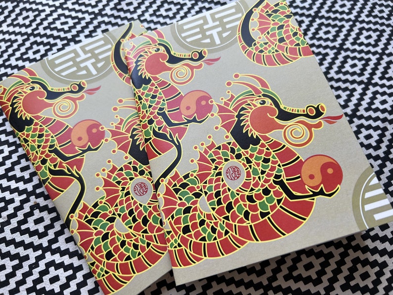 May include: Two brown notebooks with a red, green, black, and yellow dragon design. The dragon is holding a yin yang symbol. The notebooks have a gold double happiness symbol on the cover.