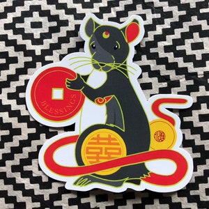 May include: A sticker featuring a stylized black rat holding a red coin with the word "Blessings". The rat has red ears and a red and gold design. A red ribbon and gold coins are also included.