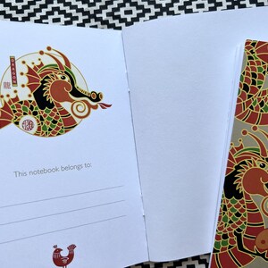 May include: A red, green, and gold dragon illustration on a white notebook cover. The dragon is holding a yin yang symbol. The text "This notebook belongs to:" is printed below the illustration.