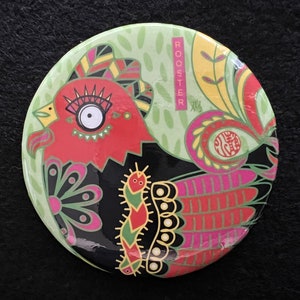 May include: A round, colorful pin with a rooster design. The rooster is red, black, and green with a large eye. The pin has the word "ROOSTER" written vertically on the right side and the Chinese character for "rooster" below it.