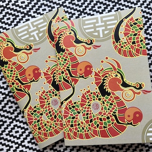 May include: Two beige notebooks with red, black, and green dragon illustrations. The dragons are holding yin-yang symbols. The notebooks have a gold Chinese character on the front.