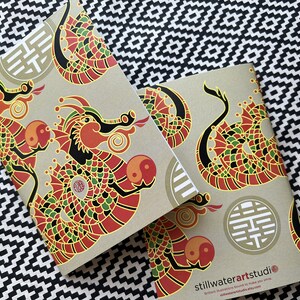 May include: Two brown notebooks with a repeating pattern of red, green, and black dragons with gold accents. The notebooks have a gold symbol on the front and the text "stillwaterartstudio" on the back.