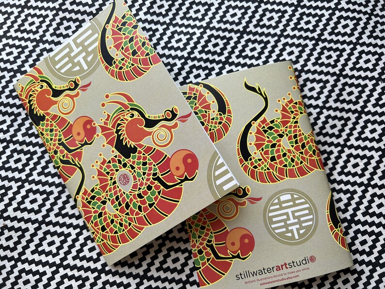 May include: Two brown notebooks with a repeating pattern of red, green, and black dragons with gold accents. The notebooks have a gold symbol on the front and the text "stillwaterartstudio" on the back.