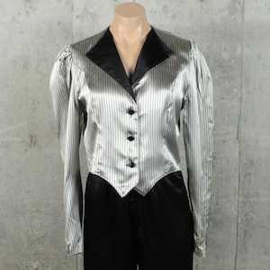 May include: A long-sleeved jacket featuring a silver and black pinstripe pattern with a solid black lapel and three black buttons down the front. The garment has a pointed hemline at the waist and gathered shoulders, presented on a mannequin.