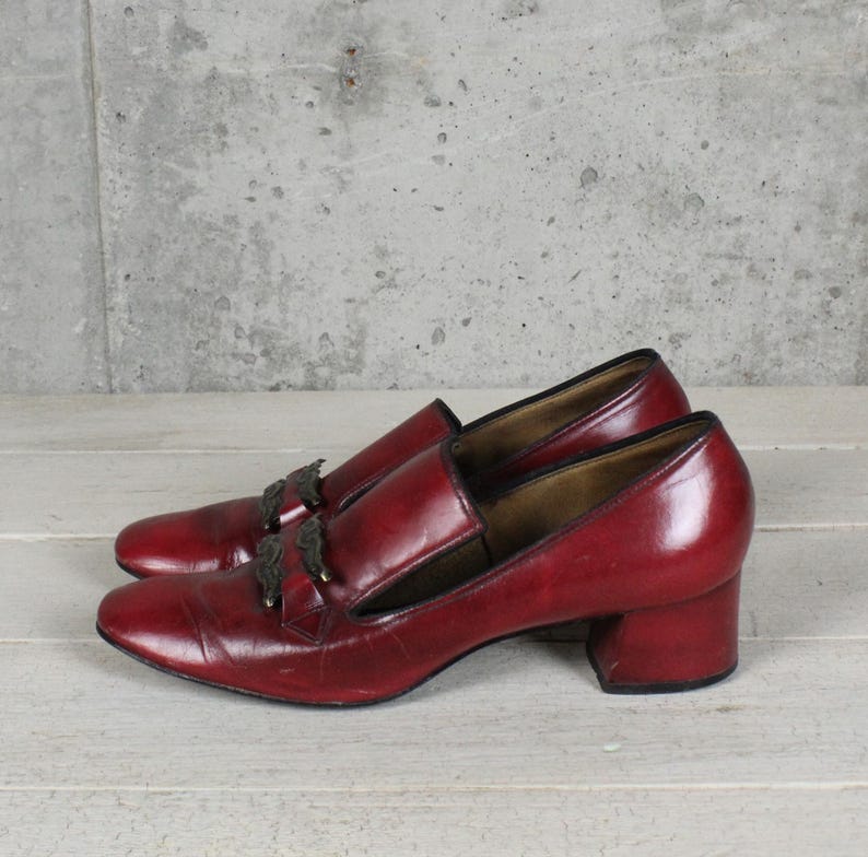 K&ouml;nnte beinhalten: Ein Paar dunkelrote Leder-Loafer mit Blockabsatz und einem dekorativen metallischen Schmuckelement auf dem Vorderblatt. Die Schuhe haben eine glatte Oberfl&auml;che, eine abgerundete Schuhspitze und ein beiges Innenfutter im klassischen Slipper-Design.