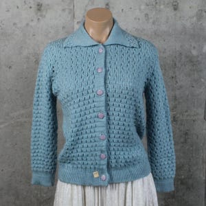 Könnte beinhalten: Hellblauer Cardigan im Vintage-Stil mit Kragen und langen Ärmeln. Der Pullover hat ein strukturiertes, offenes Strickmuster und wird mit sechs lavendelfarbenen Knöpfen geschlossen. Ein kleines Etikett ist am unteren Saum sichtbar.