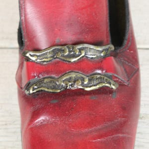 K&ouml;nnte beinhalten: Ein dunkelroter Leder-Loafer mit zwei horizontalen, verzierten Metallspangen in antikem Messington auf dem Vorderblatt. Der Schuh hat eine glatte, leicht abgenutzte Textur mit sichtbaren nat&uuml;rlichen Falten und einem dunklen Innenfutter.