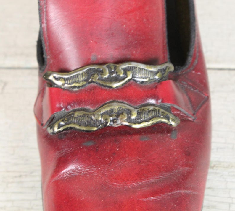 K&ouml;nnte beinhalten: Ein dunkelroter Leder-Loafer mit zwei horizontalen, verzierten Metallspangen in antikem Messington auf dem Vorderblatt. Der Schuh hat eine glatte, leicht abgenutzte Textur mit sichtbaren nat&uuml;rlichen Falten und einem dunklen Innenfutter.